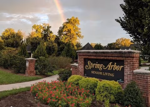 Entrance sign for Spring Arbor Senior Living made of brick with a black and gold plaque, surrounded by green bushes and red flowers, with a rainbow visible in the cloudy sky above and trees in the background.