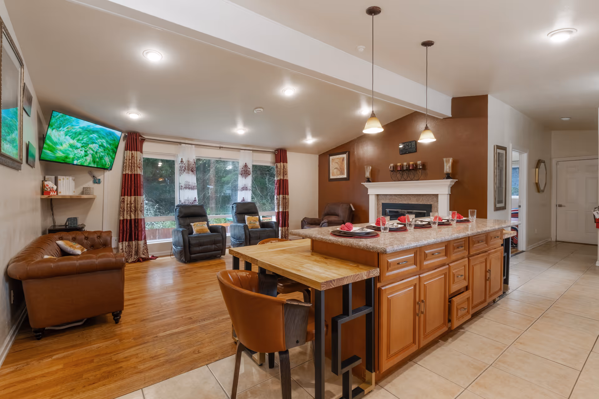 A spacious living room area with wooden flooring and a tiled kitchen island in the foreground. The kitchen island has a granite countertop with place settings including plates, glasses, and red napkins. There are two pendant lights hanging above the island. The living room features a brown leather couch, two black recliners, and a brown armchair near a white fireplace with decorative items on the mantel. Large windows with red and beige patterned curtains allow natural light to enter the room. A flat-screen TV is mounted on the wall above a small shelf with books and a phone.