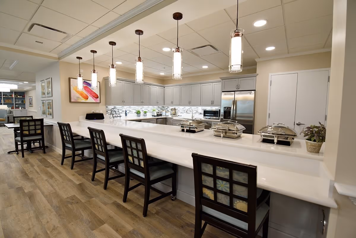A modern kitchen area with a long white countertop bar and five high chairs with patterned backs. Above the counter, there are five pendant lights hanging from the ceiling. The kitchen features light gray cabinets, a stainless steel refrigerator, a microwave, and a backsplash with a white and gray tile design. There are three silver food warmers on the counter and a small potted plant on the right side. In the background, there is a dining area with tables and chairs and colorful artwork on the walls.
