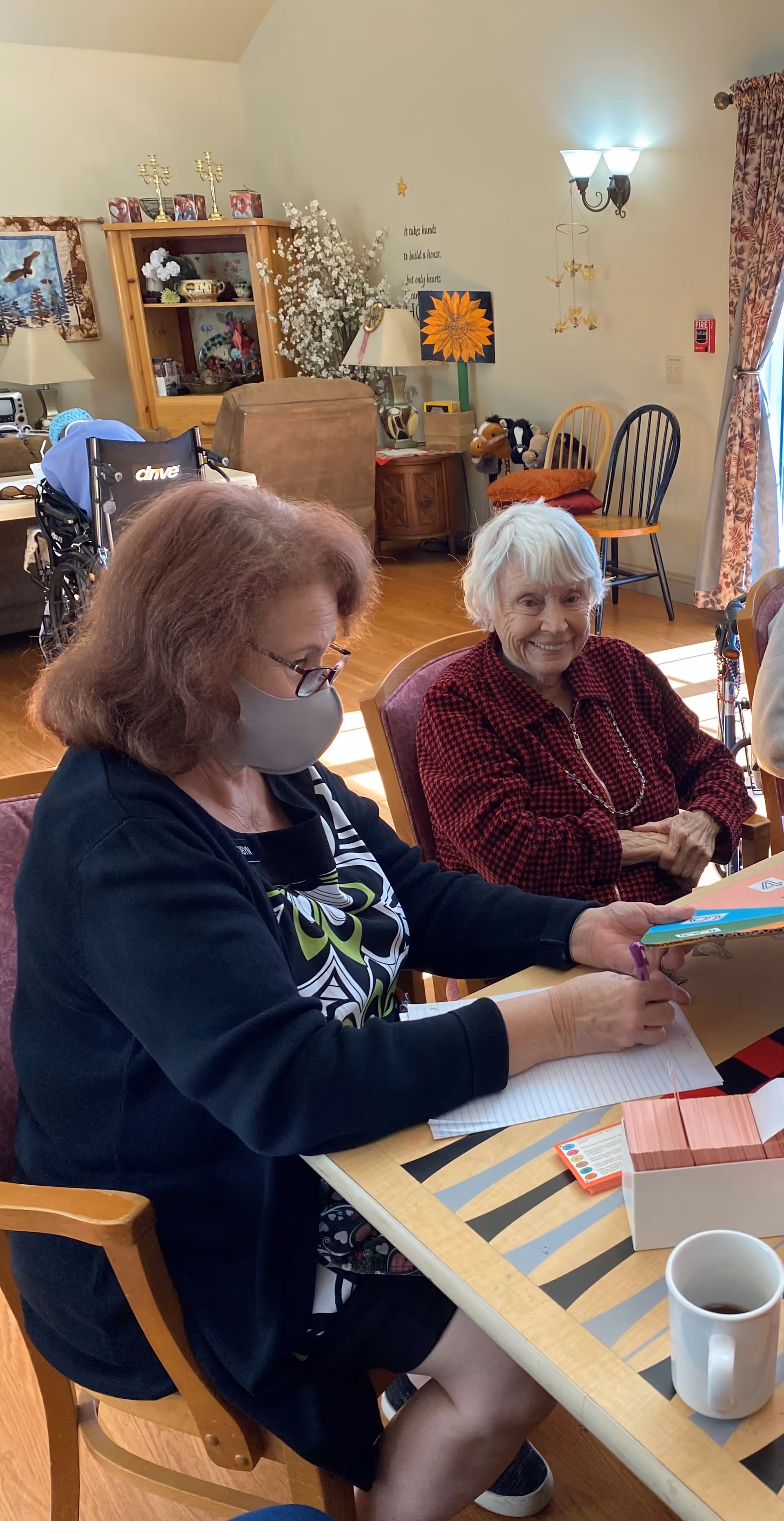 Two older women sitting at a table in a communal living room; one wearing a mask writing while the other smiles.