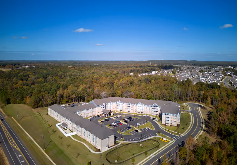 Aerial view of a large U-shaped senior living facility with a parking lot, lawns, and surrounding woods.