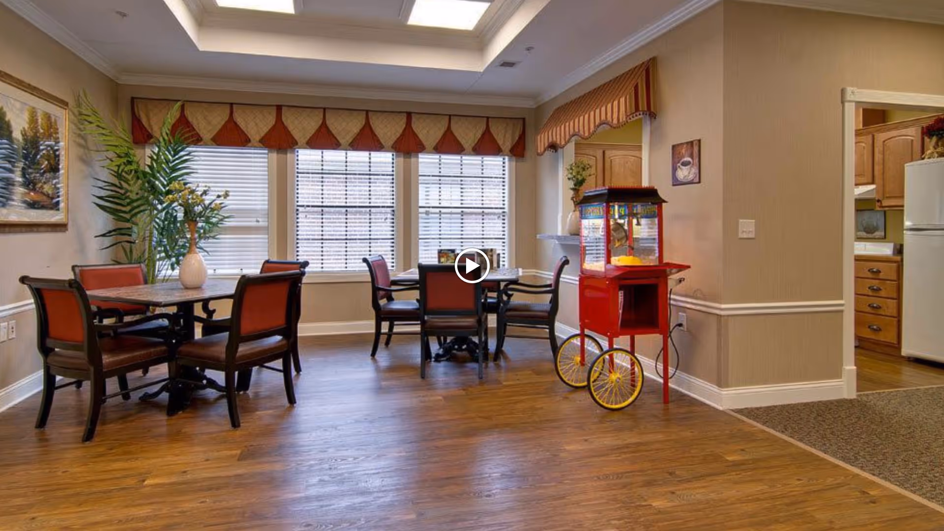 A cozy dining area in Olive Grove Terrace Senior Living featuring two tables with red cushioned chairs, a large window with blinds and valances, a decorative plant in a vase, a popcorn machine on wheels, and a view into a kitchen area with wooden cabinets and a white refrigerator.