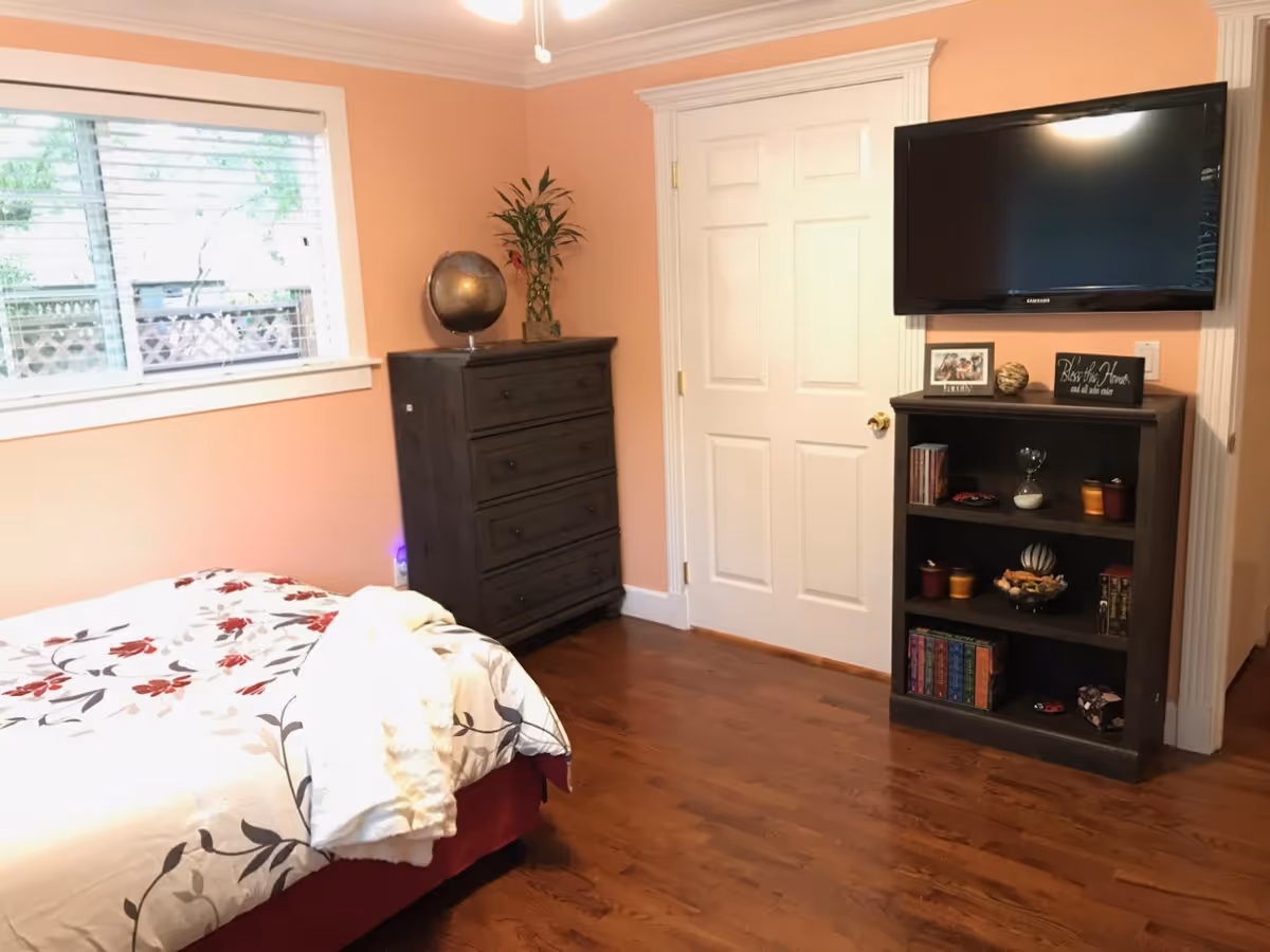 A bedroom with peach-colored walls and wooden flooring. There is a bed with a floral patterned bedspread and a white blanket draped over it. A dark wooden chest of drawers stands against the wall next to a window with white blinds. A globe and a plant are placed on top of the chest. On the adjacent wall, a flat-screen TV is mounted above a dark wooden bookshelf holding books, candles, an hourglass, and decorative items. A white door with gold-colored hinges and knob is also visible.