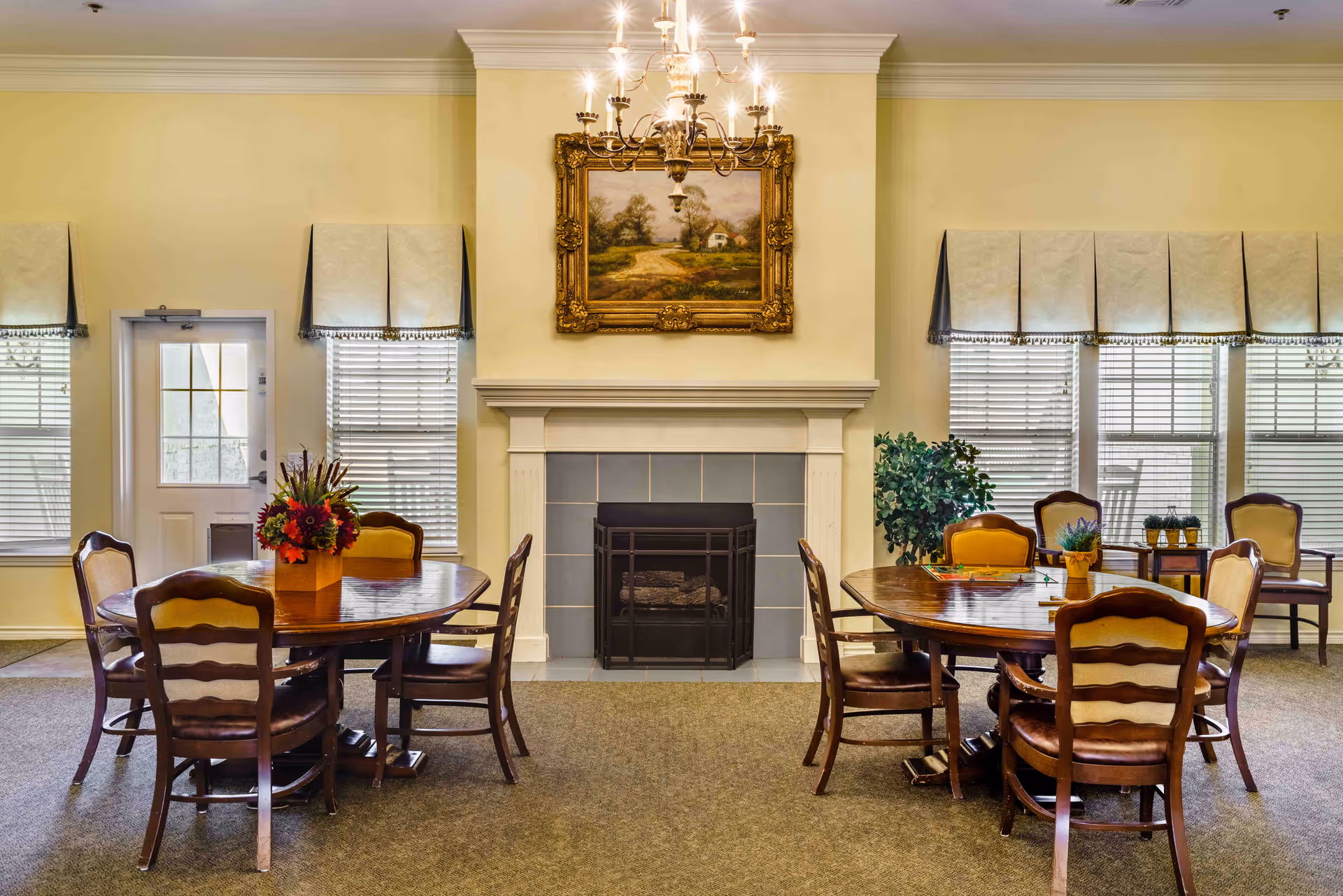 Bright communal dining room with two round wooden tables and chairs arranged around a central fireplace topped by a framed landscape painting.