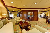 A spacious and well-lit living room area in a senior living facility with comfortable sofas, armchairs, a wooden coffee table with a decorative vase, patterned carpet, and a wooden partition with a painting. The room has warm lighting and a cozy atmosphere.