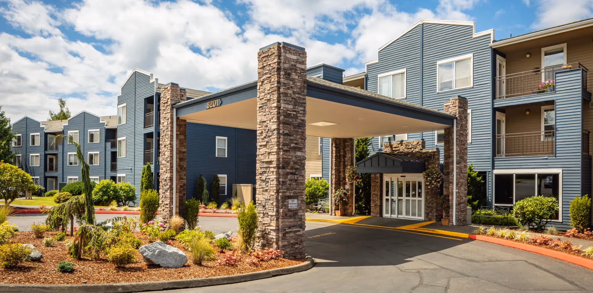Exterior view of Cogir at The Narrows senior living facility showing a covered entrance supported by stone pillars, landscaped garden beds with shrubs and rocks, and a multi-story building with blue siding and balconies under a partly cloudy sky.