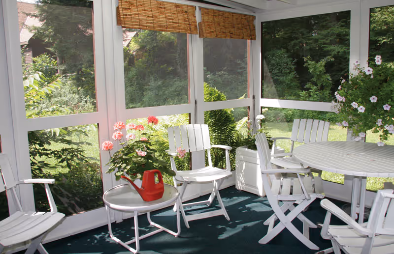 A bright screened sunroom with white wooden chairs and table, potted flowers, and a red watering can on a small table.