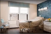 A bright and clean patient room featuring a hospital bed with a beige quilted cover, a small overbed table, a white dresser with a vase of yellow flowers, two chairs near the window, and a blue accent wall behind the bed. The room has two windows with blinds and an air conditioning unit installed in one window.