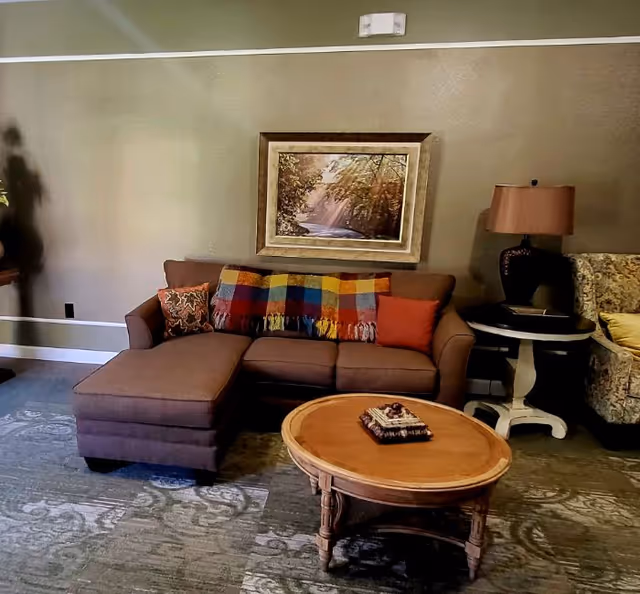 Cozy living room with a brown sectional sofa adorned with a colorful throw, a round wooden coffee table, side table with lamp, and framed landscape painting on the wall.