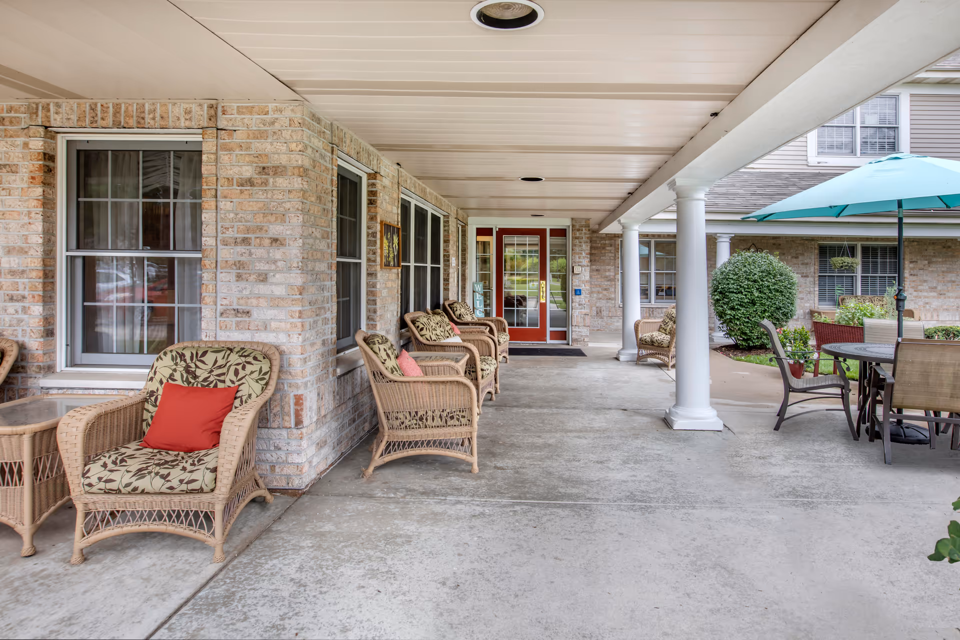 Covered outdoor patio area at a senior living facility with wicker chairs featuring floral cushions and red pillows, a round table with chairs and a teal umbrella, brick walls, white columns, and glass doors at the entrance.
