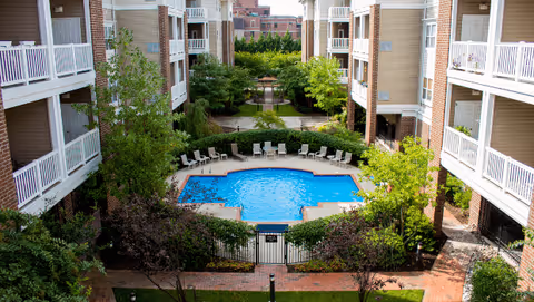 View of a rectangular outdoor swimming pool surrounded by lounge chairs and greenery, situated in the courtyard of a multi-story residential building with balconies overlooking the pool area.