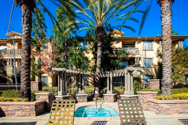 Outdoor courtyard with a small circular spa, a pergola, lounge chairs, palm trees, and a three-story apartment building in the background.