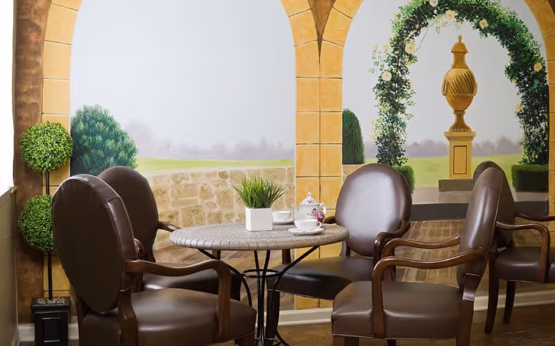 A cozy indoor seating area with four brown leather chairs arranged around a round table. The table has a small potted plant and a tea set on it. The background features a mural painted to resemble an outdoor garden scene with stone arches, greenery, and a decorative urn under an archway.