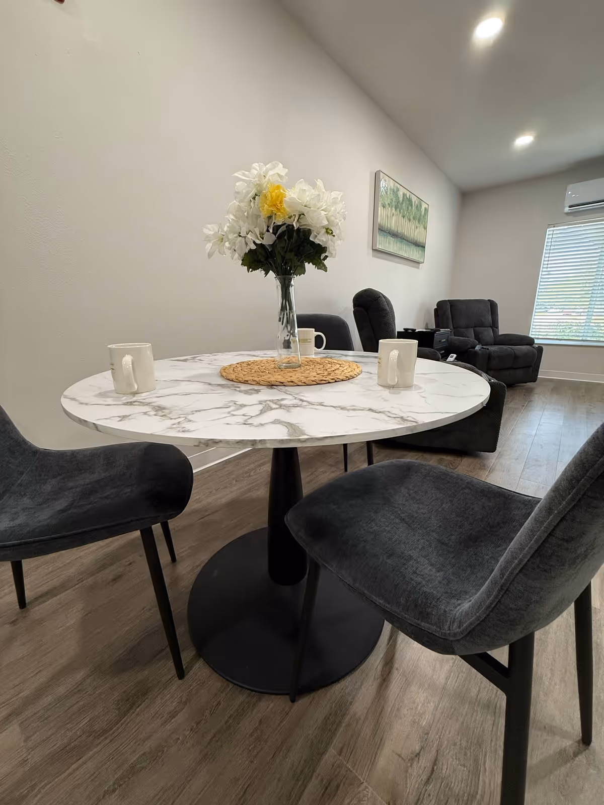 A modern living area with a round marble-top table surrounded by four dark gray upholstered chairs. On the table, there is a vase with white and yellow flowers and three white mugs. In the background, there are two dark gray recliner chairs, a window with blinds, a wall-mounted air conditioner, and a framed landscape painting on the wall.