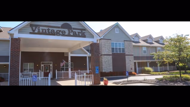 Front exterior view of the Vintage Park at Stanley senior living facility showing the entrance with a covered drop-off area, brick and siding facade, multiple windows, and a small landscaped area with a tree and plants.