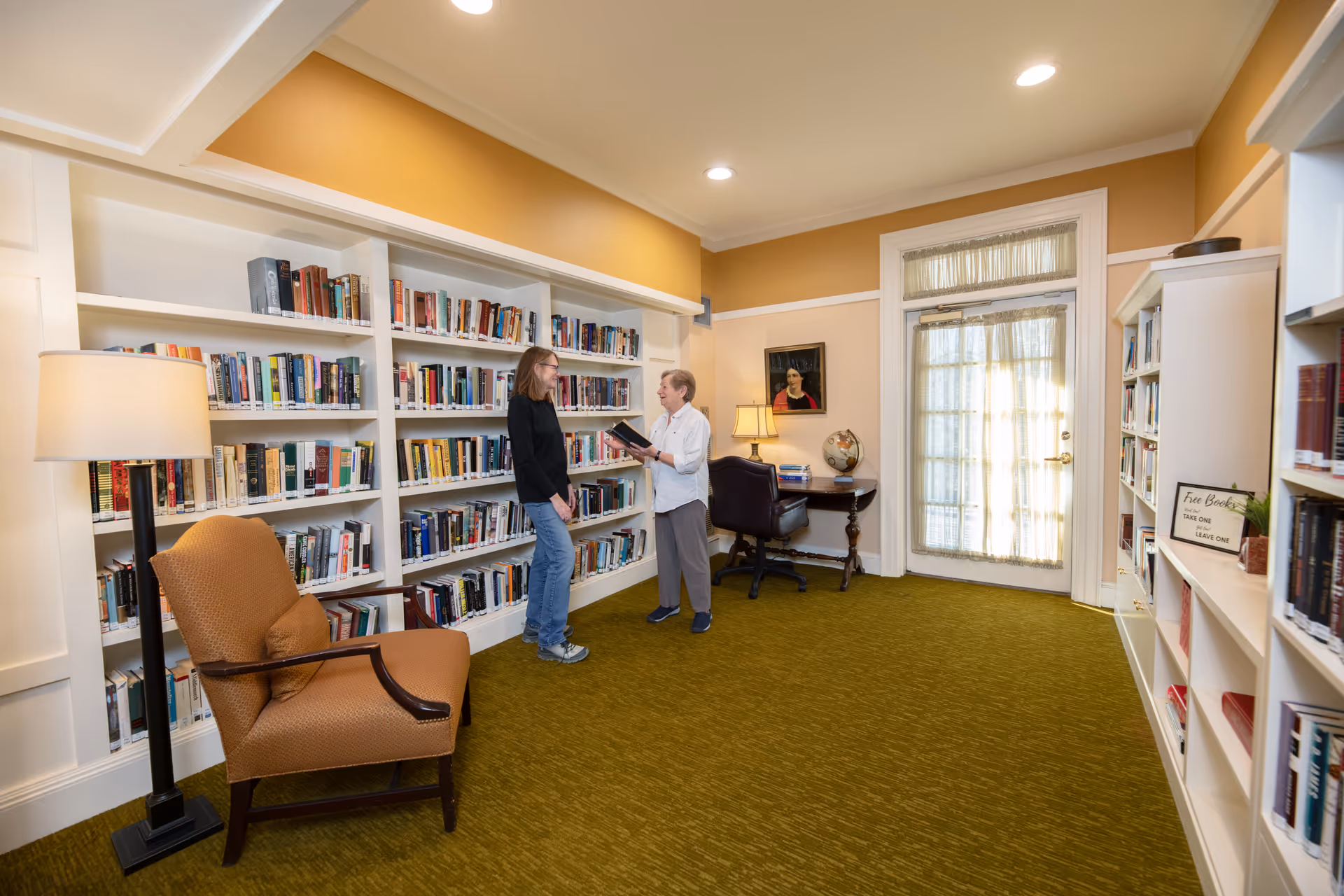 A cozy library room with bookshelves filled with books along the walls. Two women are standing and talking near the shelves, one holding a book. The room has a green carpet, a comfortable armchair with a floor lamp beside it, a small desk with a globe and lamp, and a door with sheer curtains letting in natural light.