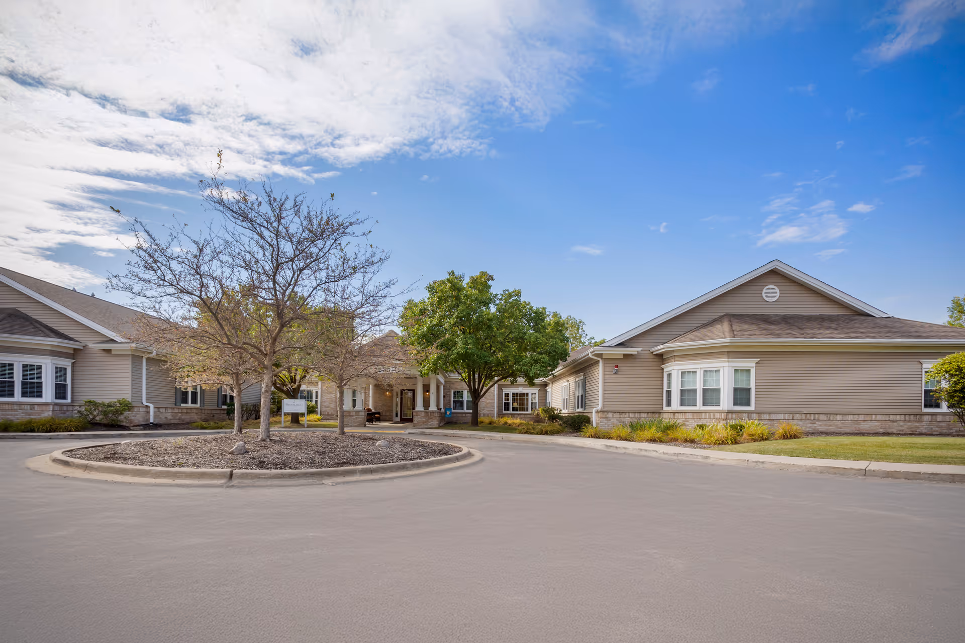 Exterior view of Brookdale Delta facility showing a single-story building with beige siding and a circular driveway with a landscaped island featuring trees and shrubs under a partly cloudy blue sky.