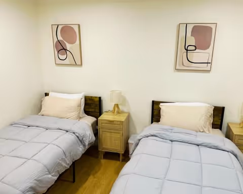 A bedroom with two single beds separated by a wooden nightstand with a lamp. Each bed has a light gray comforter and a beige pillow. Two abstract art pieces hang on the white wall above the beds.