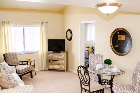 Sunlit living room with a sofa and armchairs, a small TV on a cabinet, and a round glass dining table with place settings.