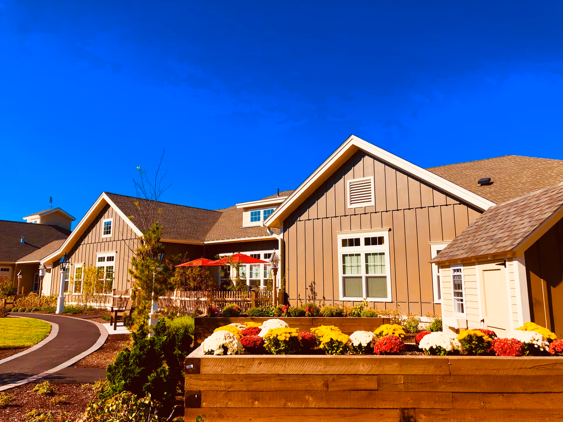 Exterior view of a senior living facility building with beige siding and brown shingled roof under a clear blue sky. In the foreground, there is a raised wooden flower bed filled with colorful flowers including white, yellow, and red blooms. A paved walkway curves along the left side with benches and small trees lining the path. Red umbrellas are visible near the building, suggesting an outdoor seating area.
