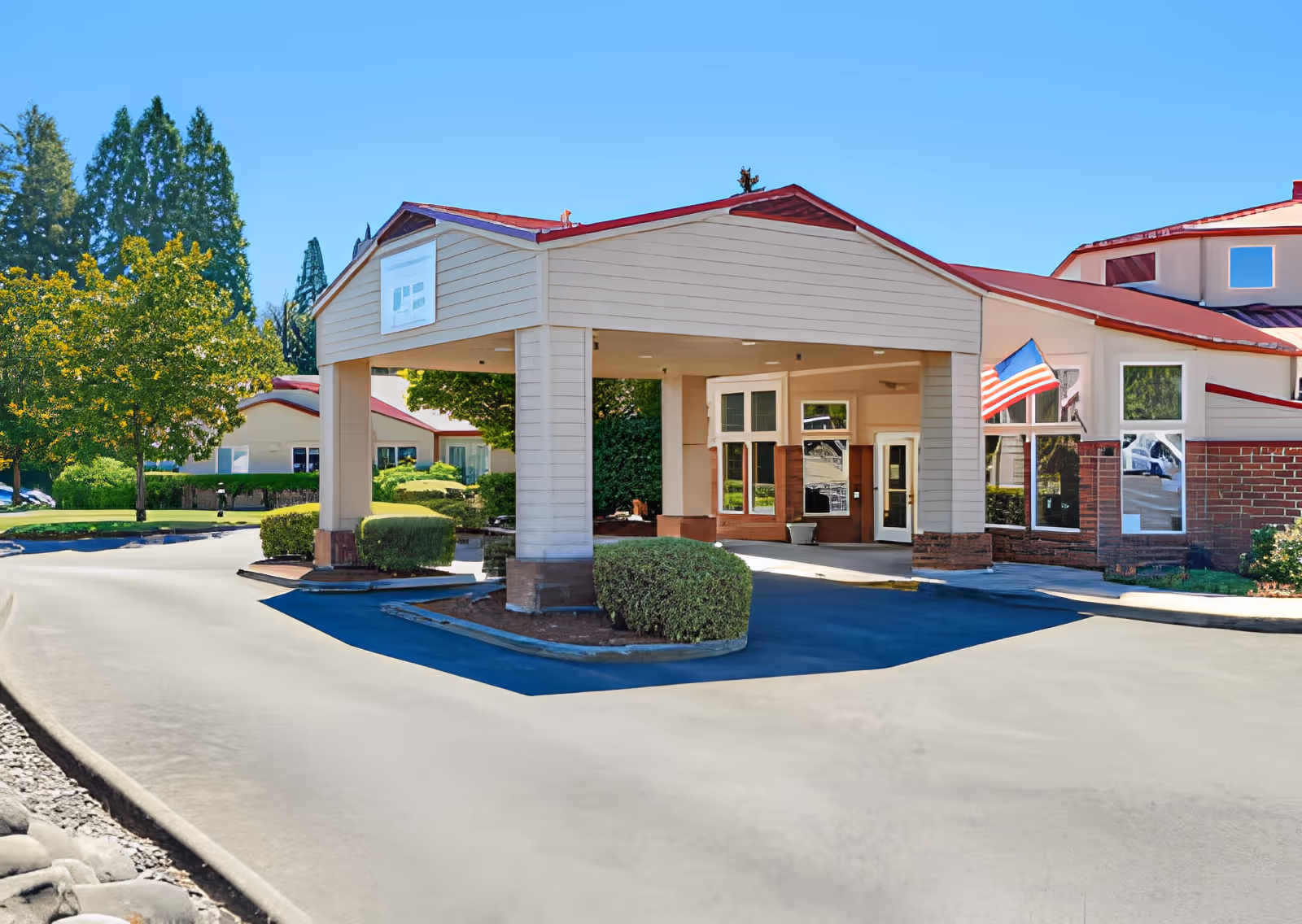 Exterior view of Elliott Residence showing the main entrance with a covered driveway, surrounded by well-maintained bushes and trees under a clear blue sky.