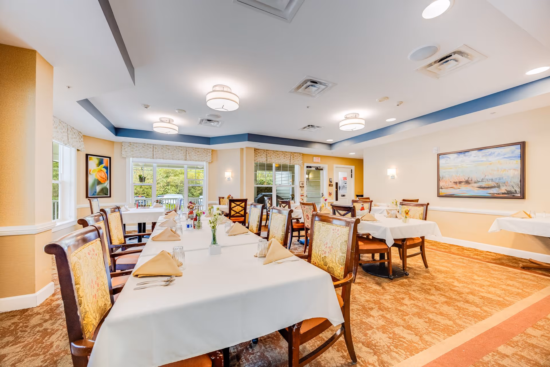 A bright and spacious dining room with multiple tables covered in white tablecloths and set with beige napkins, glasses, and silverware. The room features upholstered wooden chairs, large windows letting in natural light, framed artwork on the walls, and ceiling lights. The carpet is patterned in warm tones, and the walls are painted in soft beige with white trim.