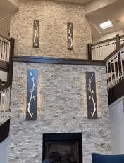 Interior view of a living room area with a tall stone fireplace wall featuring four vertical decorative panels with branch designs. The room has a high ceiling with stair railings visible on both sides and a skylight above.