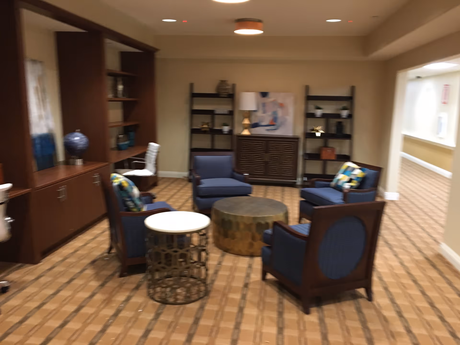A cozy sitting area with four blue cushioned armchairs arranged around two round tables, one metallic and one white. The room has wooden shelves and cabinets along the walls, decorated with vases, books, and small plants. The floor is carpeted with a patterned design, and the walls are painted beige. There is a hallway visible to the right.