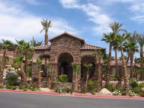 Exterior view of a senior living facility building with stone and stucco facade, arched entryways, and a tiled roof. The building is surrounded by palm trees and desert landscaping under a partly cloudy blue sky.
