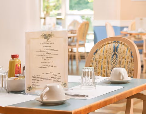 A dining table set with white cups and saucers, upside-down drinking glasses, and a menu card displaying lunch options. The background shows more tables and chairs in a softly lit dining room with large windows letting in natural light.