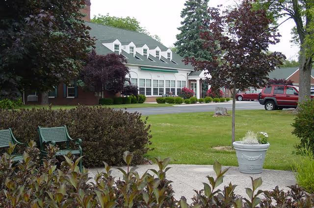 Exterior view of a senior living facility with a brick building, green roof, and multiple windows. The foreground features a landscaped garden with bushes, trees, a white planter with flowers, and green benches. A red SUV is parked near the building.