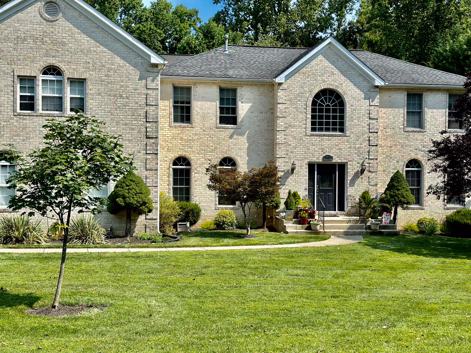 Exterior view of a two-story brick building with multiple windows and a central entrance door. The building is surrounded by a well-maintained lawn, small trees, shrubs, and potted plants near the entrance. The sky is clear and the scene is bright and sunny.