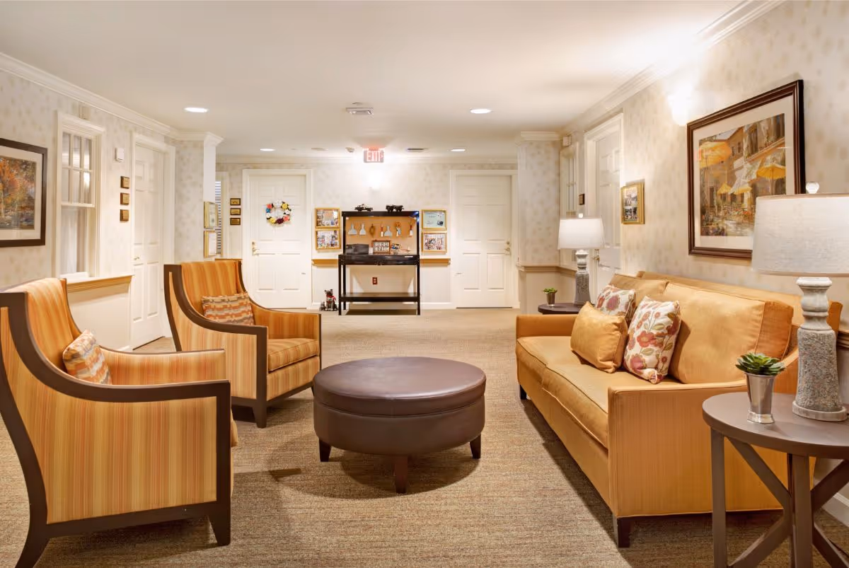 A cozy sitting area in a senior living facility with a mustard yellow sofa adorned with floral and solid pillows, two striped armchairs, a round brown ottoman in the center, and two side tables with lamps and small plants. The walls are decorated with framed artwork and soft lighting, and there are multiple white doors and a bulletin board in the background.
