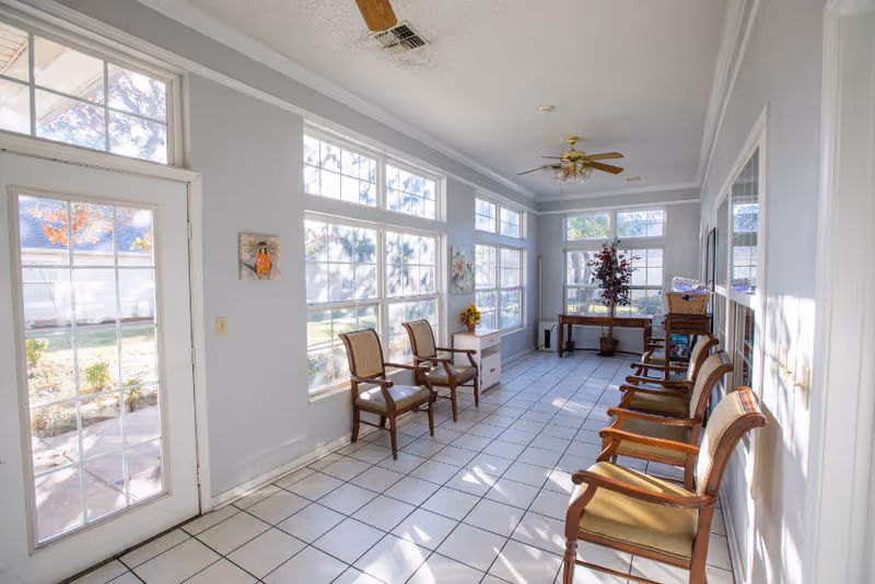 A bright, sunlit room with large windows on two sides, featuring several wooden chairs with cushions arranged along the walls. The room has white tiled flooring, light gray walls, a ceiling fan with lights, and a door leading outside. There are decorative items including a small table with a flower vase and wall art.