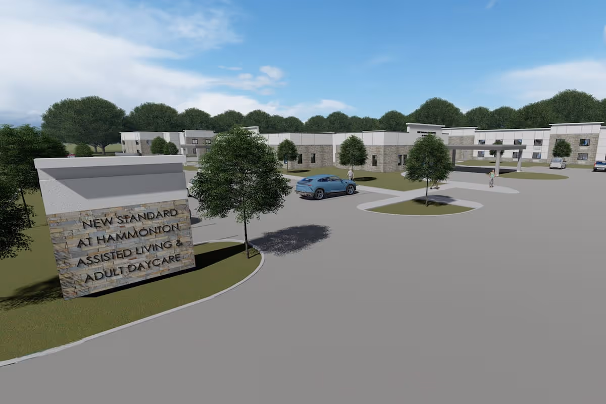 Exterior view of New Standard Senior Living at Hammonton facility showing a large sign with the facility name, a parking area with cars, trees, and a two-story building under a partly cloudy sky.