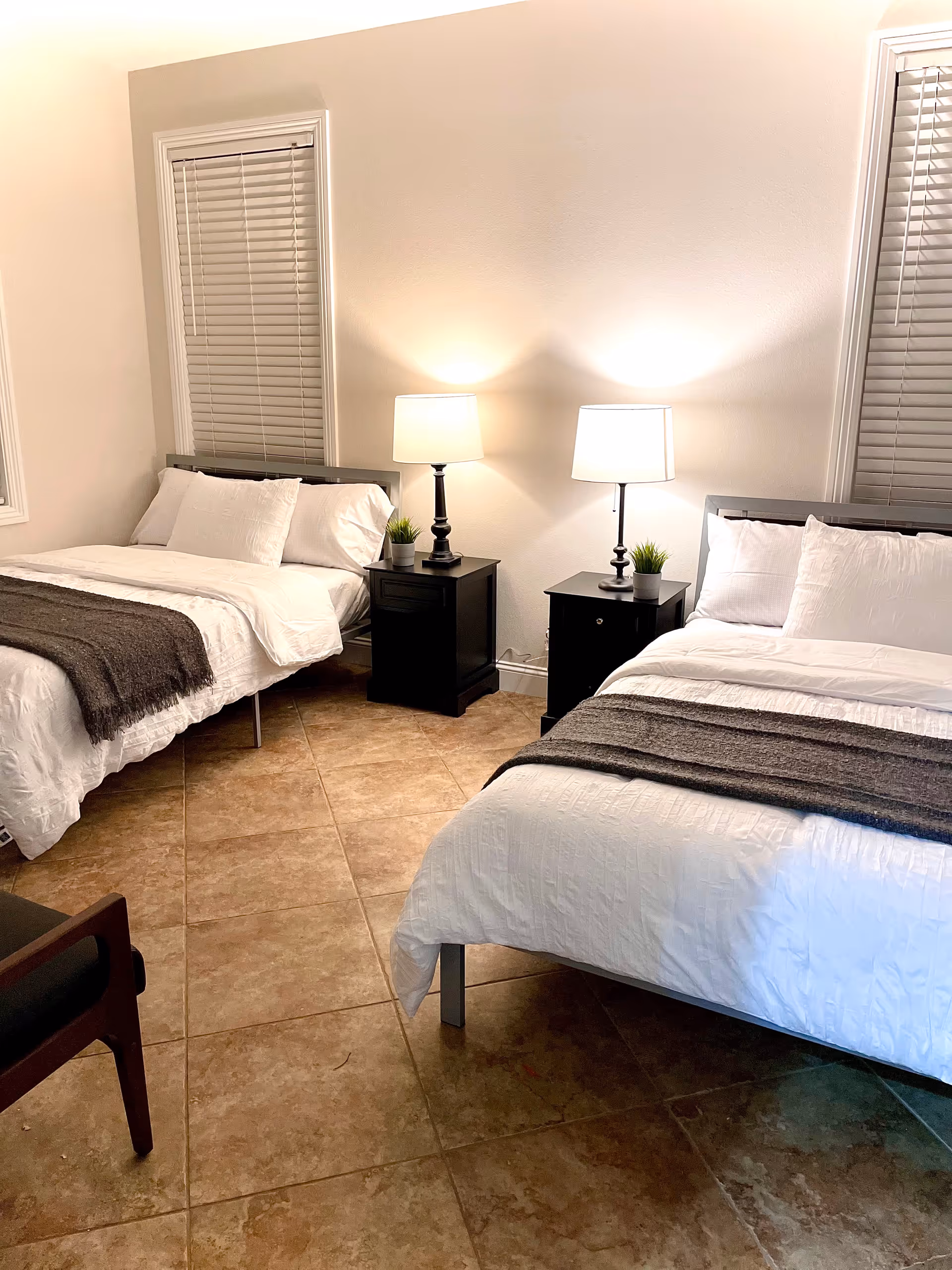 A bedroom with two beds, each with white bedding and a dark gray throw blanket. Between the beds are two black nightstands, each with a lamp and a small green plant. The room has tiled flooring and two windows with closed blinds.