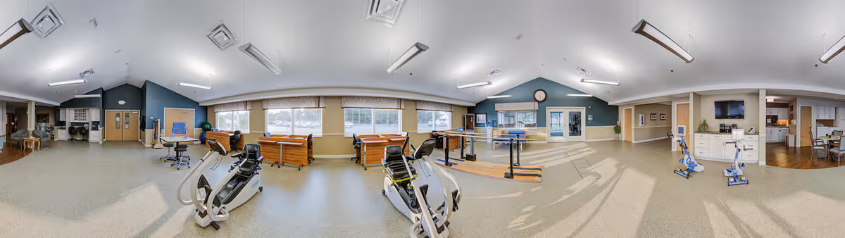 A spacious rehabilitation or therapy room in a senior living facility with exercise equipment including stationary bikes, parallel bars for walking practice, therapy tables, and a ping pong table. The room has large windows allowing natural light, light-colored flooring, and walls painted in soft blue and beige tones. There are also chairs and tables in the background, along with a clock and a television mounted on the wall.