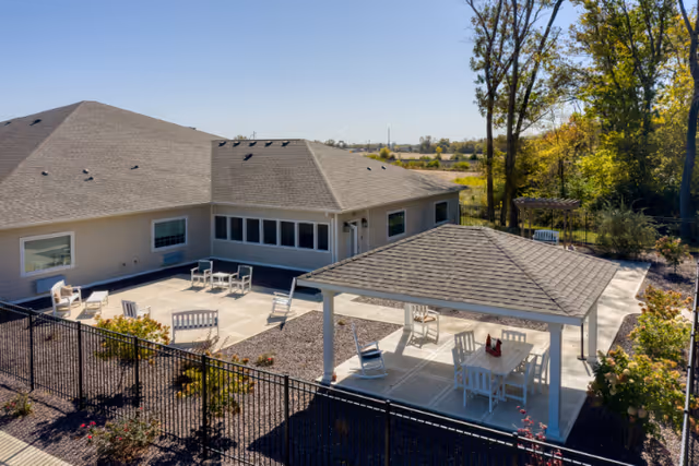 Outdoor patio area at a senior living facility with a covered seating area featuring a table and chairs, additional chairs and benches on the open patio, surrounded by a black metal fence and landscaping with trees and shrubs.