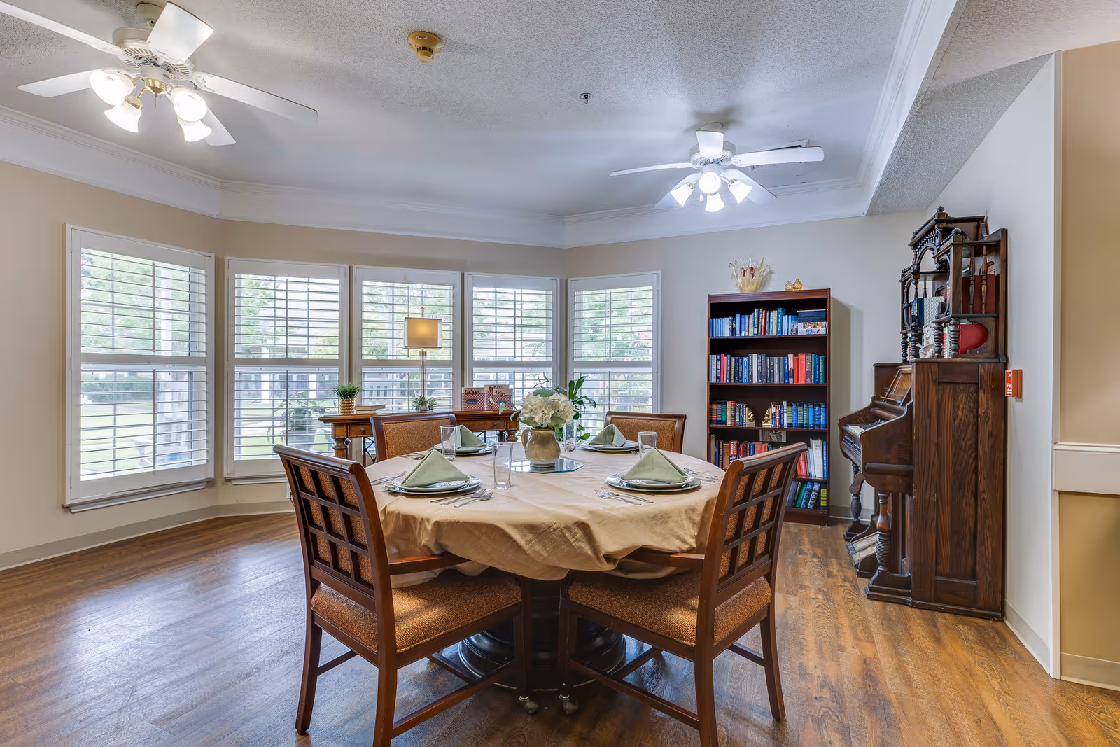 A cozy dining area with a round table set for four, featuring green napkins, plates, and glasses. The room has large windows with white shutters letting in natural light. There are two ceiling fans with lights, a wooden bookshelf filled with books, and a vintage wooden piano against the wall. The floor is wooden, and a small lamp and plants are placed on a side table near the windows.