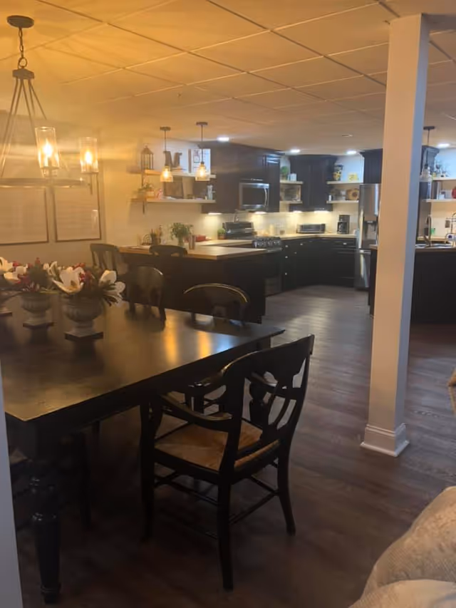 Interior view of a dining area and kitchen in Magnolia Assisted Living. The dining area features a dark wooden table with matching chairs and floral centerpieces. The kitchen has dark cabinetry, stainless steel appliances, and pendant lighting above the counter. The space has warm lighting and wooden flooring.