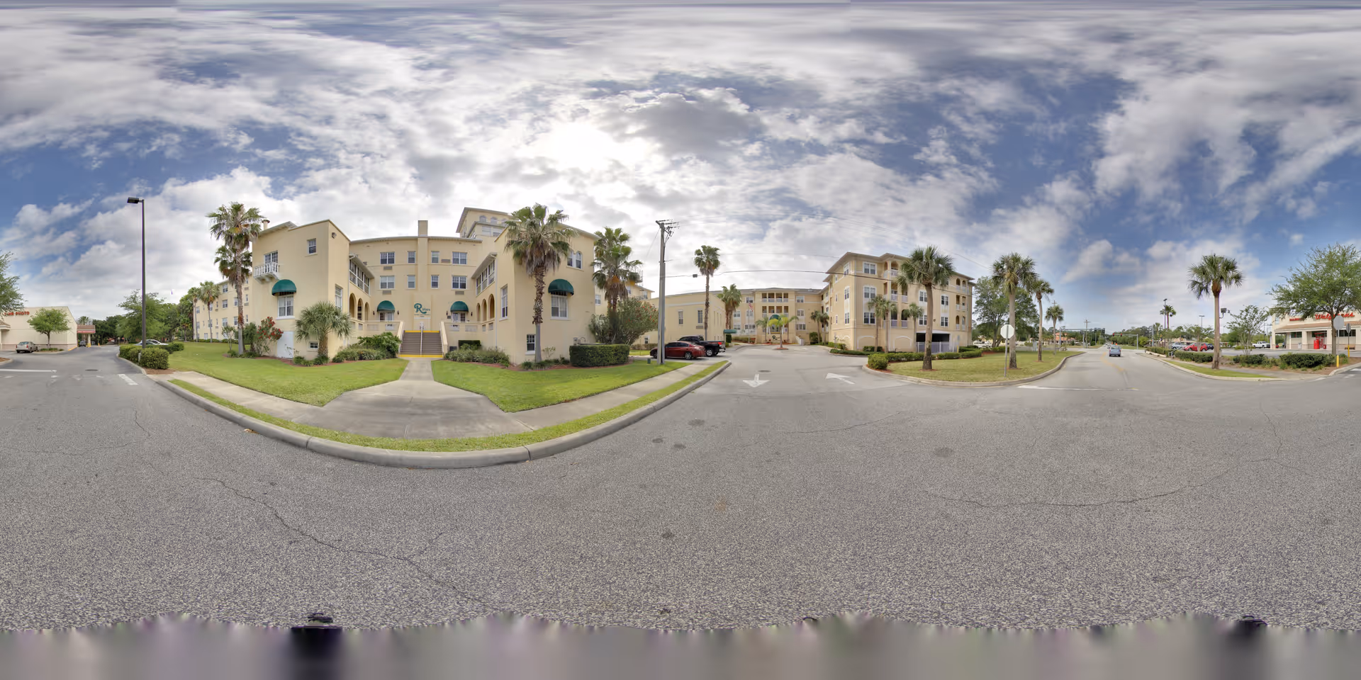 Front exterior view of a multi-story cream-colored senior living building with palm trees, lawns, and a parking area under a cloudy sky.