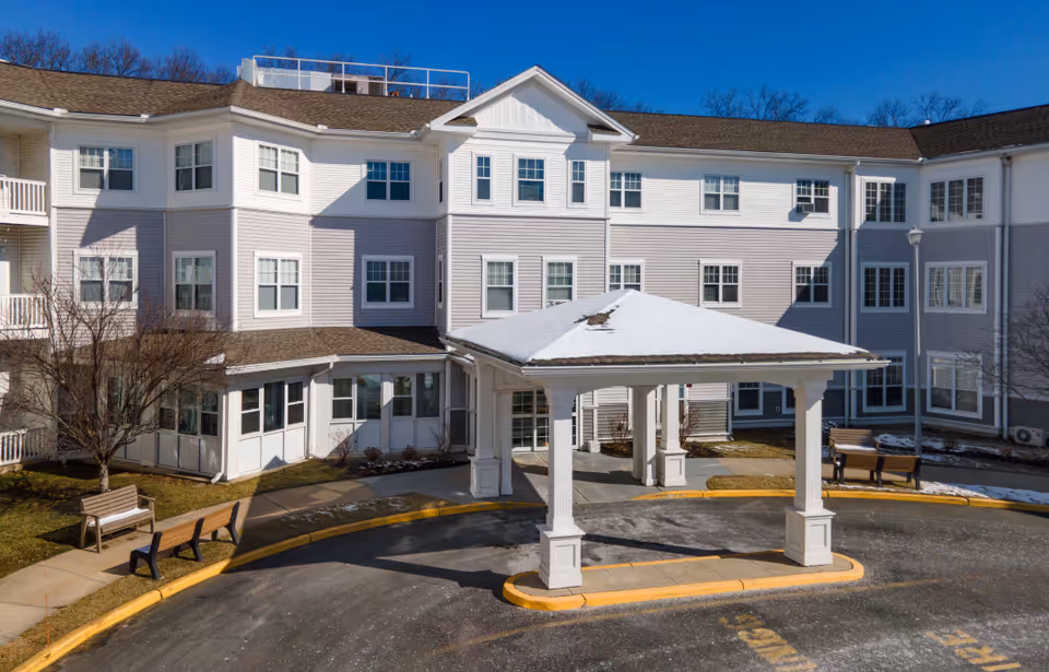 Front entrance of a multi-story senior living building with a covered porte-cochère, benches, and a circular driveway.