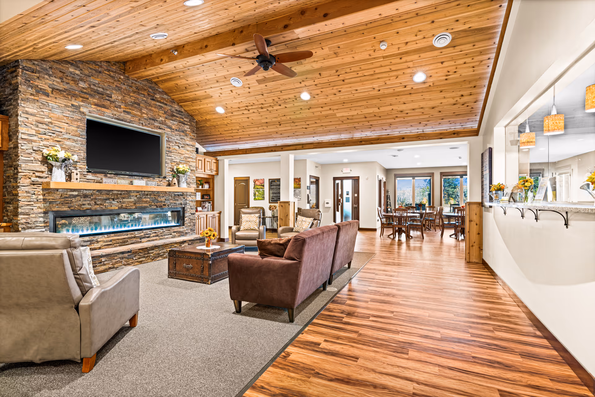 Open communal living room with a stone fireplace and TV, sofas and chairs, a wood-paneled vaulted ceiling, and a dining area visible in the background.