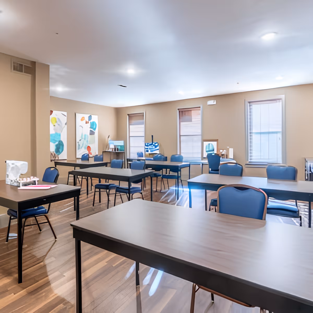 A bright room with multiple rectangular tables and blue chairs arranged in rows, featuring wooden flooring, beige walls, three windows with blinds, and abstract artwork on the walls.