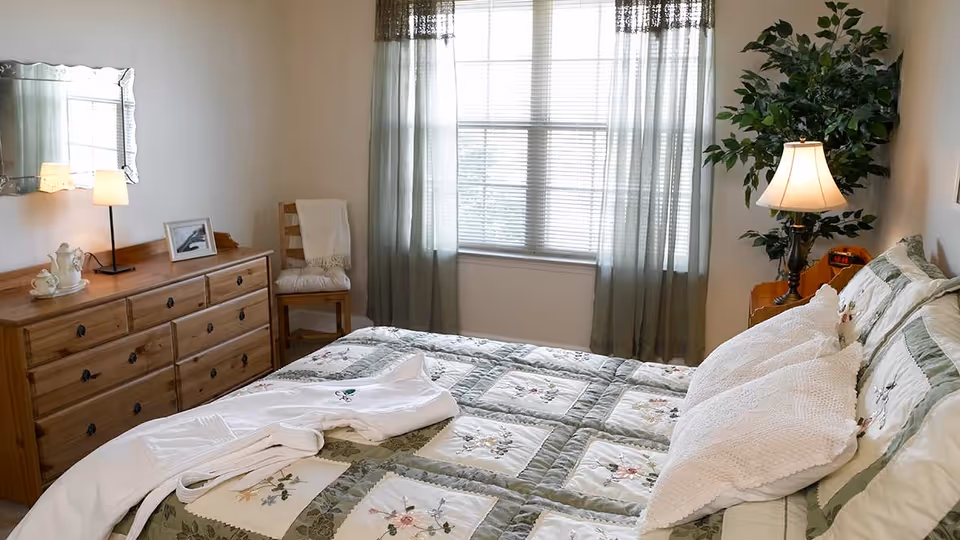 A cozy bedroom with a bed covered in a floral quilt and white pillows. A white robe is laid out on the bed. There is a wooden dresser with a lamp, a framed photo, and a tea set. A wooden chair with a white cushion and blanket is next to the dresser. The window has sheer green curtains, and a bedside table with a lamp and a potted plant is visible.