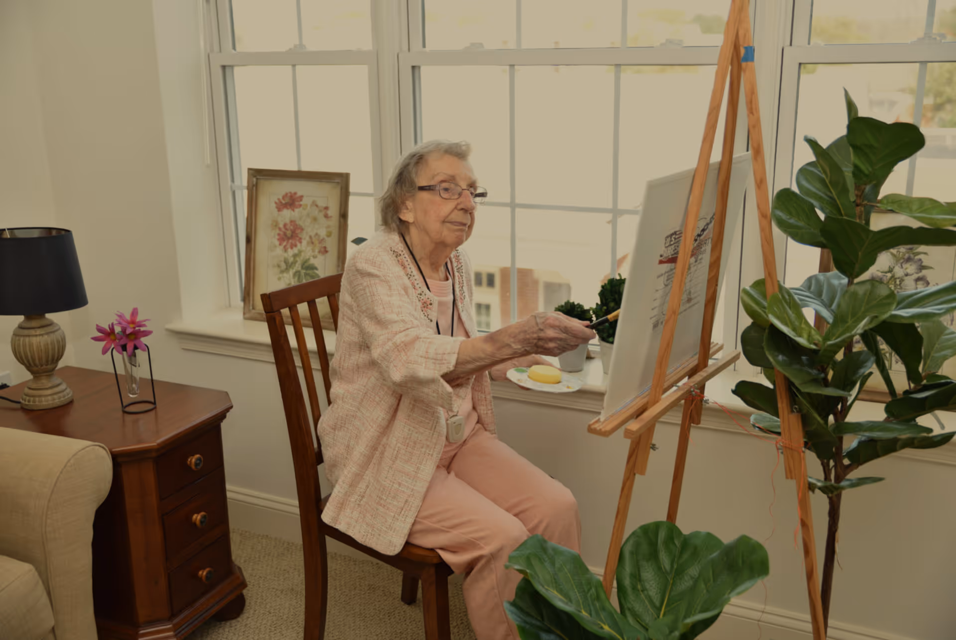 An elderly woman sitting on a wooden chair in a well-lit room, painting on a canvas set on an easel. She is wearing glasses, a light pink jacket, and pink pants. The room has large windows, a wooden side table with a lamp and a small flower vase, and several green plants nearby.