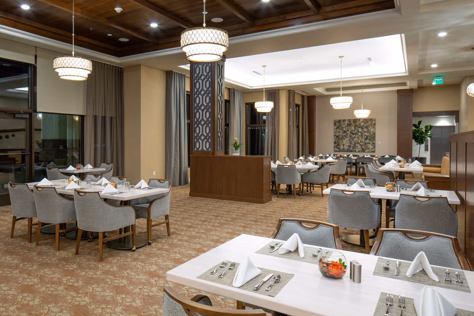 A spacious and well-lit dining room with multiple tables set for meals. Each table has neatly folded white napkins, silverware, and small floral centerpieces. The room features carpeted floors, large windows with curtains, modern hanging light fixtures, and a decorative wooden partition in the center.