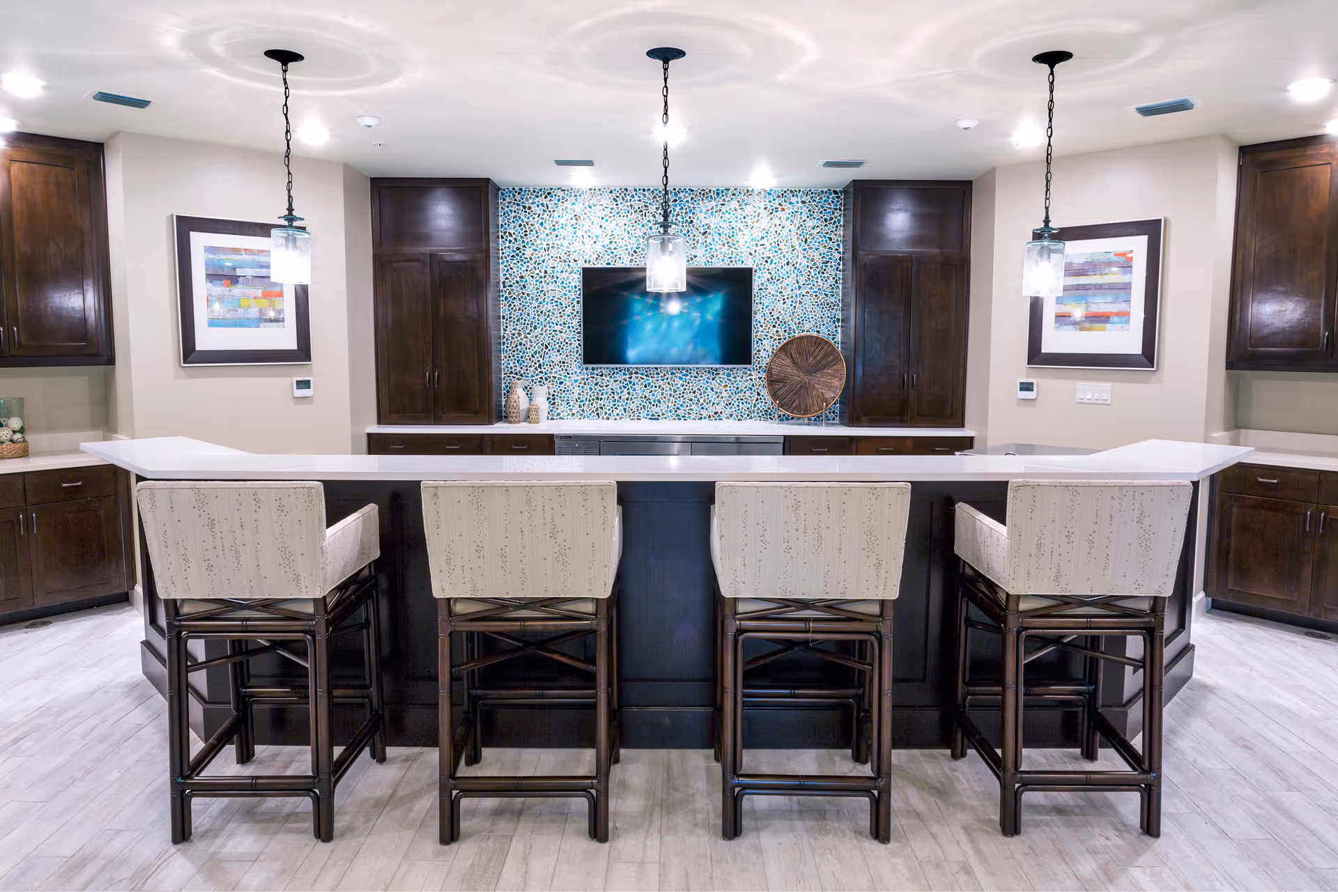 Modern communal bar area with four upholstered stools at a white countertop facing a mosaic-tiled wall with a mounted TV.