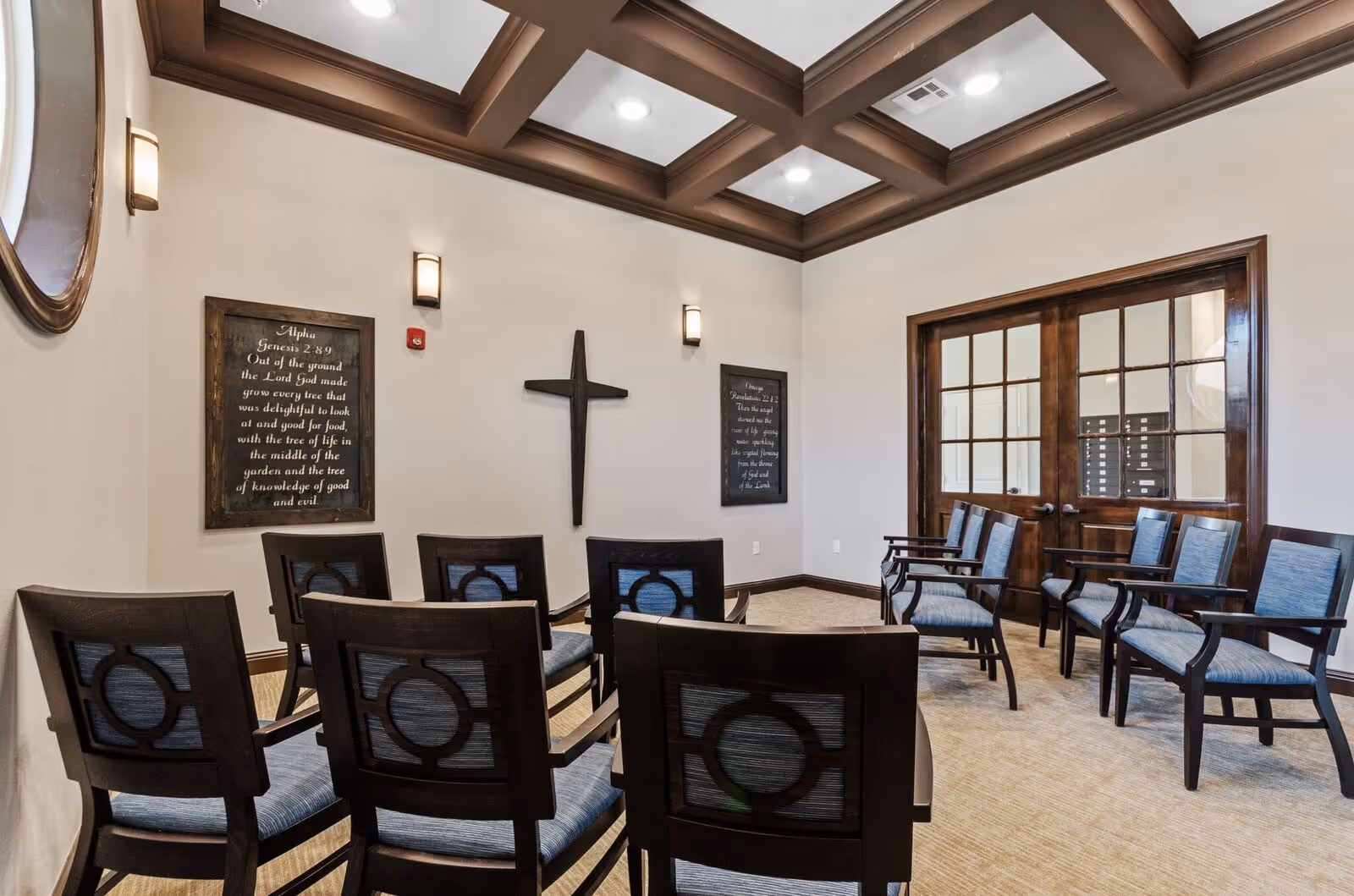 A small chapel-style interior with rows of wooden chairs facing a wall-mounted cross and framed scripture plaques, under a coffered ceiling and near double doors.