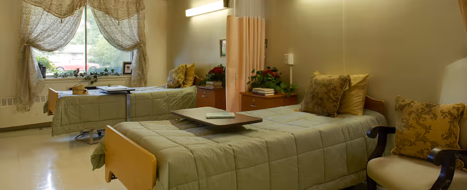 A cozy room with two single beds covered in light green quilts, each with decorative pillows. A small table is placed on one bed, and there are bedside tables with books and plants. A large window with sheer curtains lets in natural light, and an upholstered chair with a patterned cushion is visible on the right.